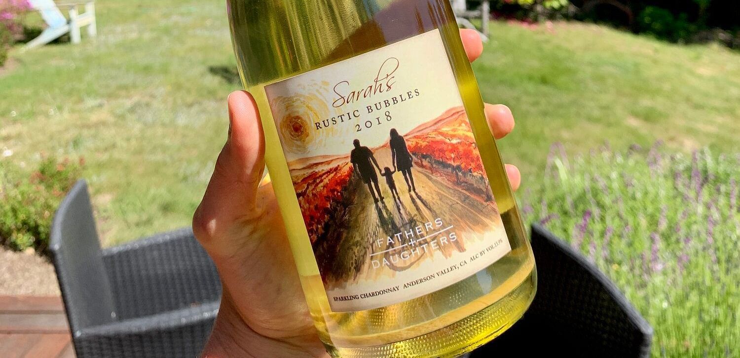 A hand holding a bottle of Sarah's Rustic Bubbles sparkling wine outdoors.
