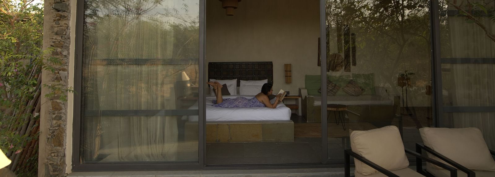An image of a woman lying on the bed where an expansive glass door leads to the outdoor seating area - Utsav Camp Sariska