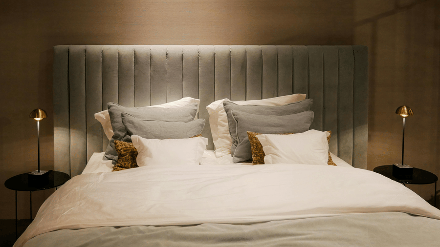 A close-up of a bed with a light grey headboard, white linens, and multiple accent pillows and 2 small black bedside tables hold slim lamps.