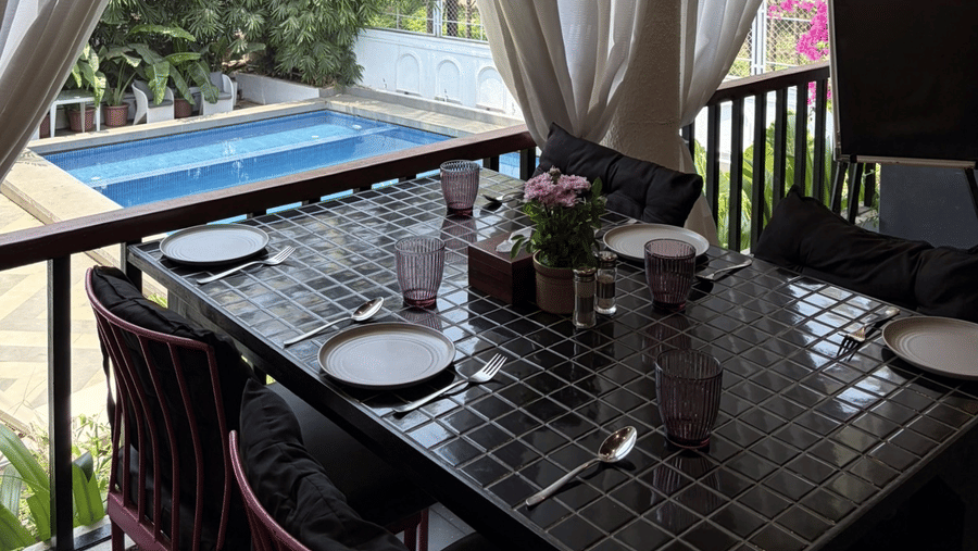 A set dining table stands on a balcony with white curtains, railings, and a view of a swimming pool at Bougainvillea, Alibaug.