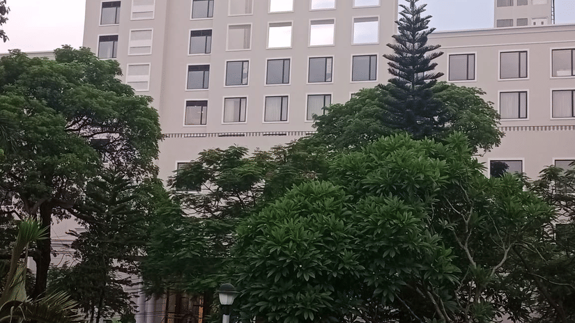 Facade of Polo Hotel, Agartala with rows of square windows, decorative banding and lush frangipani and pine trees in the foreground.