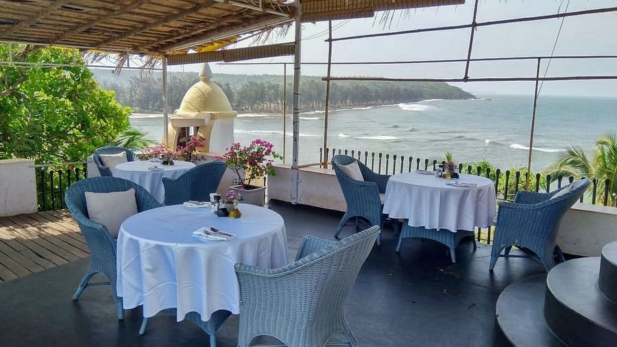 An outdoor dining area at Fort Tiracol, Goa, with tables set with white tablecloths and light blue chairs, under a shaded canopy. 