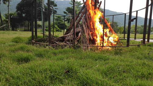 Large bonfire burning in the middle of a grassy field with trees and mountains in the background, set for an outdoor event - Black Thunder, Coimbatore