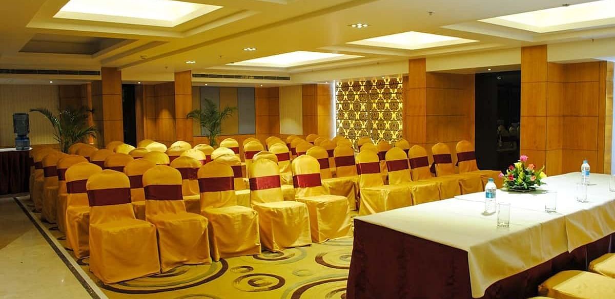 The image shows a conference room with rows of chairs facing a table with water bottles and flowers - Lords Eco Inn Navsari.