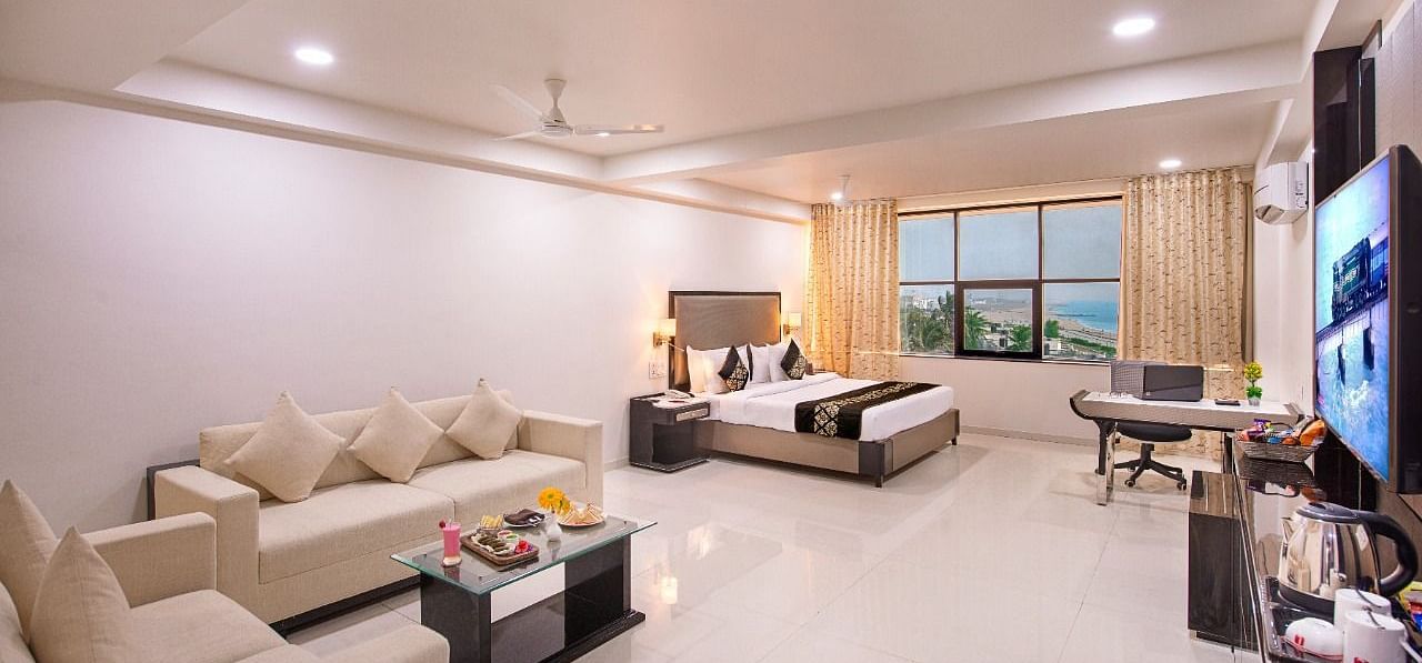 A spacious hotel suite at Lords Inn Porbandar, offers a seating area with sofas, a dining table, and a view through large windows.