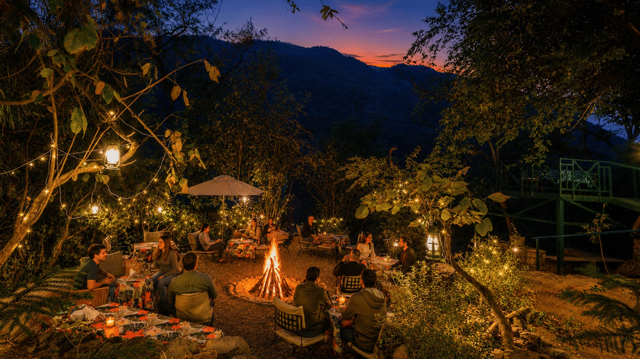 Guests gathering around a glowing central bonfire under fairy lights at dusk at Neemrana's Glasshouse on The Ganges - 21st Century, Rishikesh.