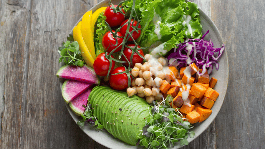 Overhead view of a salad bowl with sliced avocado, cherry tomatoes on the vine, chickpeas, roasted sweet potato cubes, red cabbage, and microgreens.