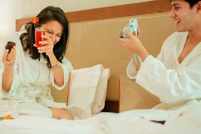 Two people sitting on a bed in robes, smiling and taking pictures with vintage-style instant cameras at Karma Chalets.