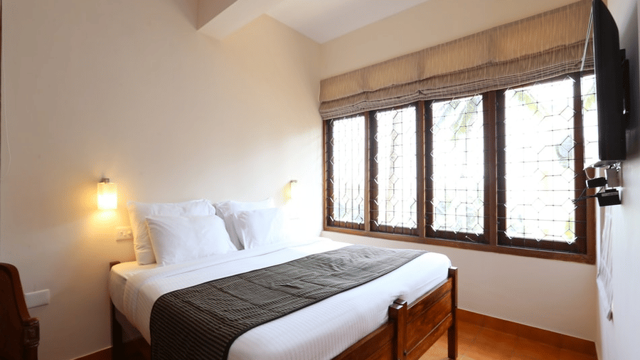 A deluxe room bedroom at Abad Serviced Villas, Kovalam with a double bed, bedside table, lamp, and large windows.