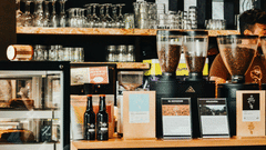 A coffee shop showing the counter with espresso machines, coffee beans, glassware, a display case, and a wooden table in the foreground.
