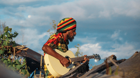 A Rastafarian playing guitar while wearing a beanie with the Jamaican flag colours inside Rastafari indigenous village.