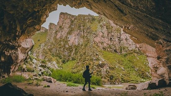 An image of a man in standing on the entrance of the cave which is surrounded by a rocky hill