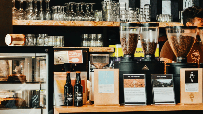 A coffee shop showing the counter with espresso machines, coffee beans, glassware, a display case, and a wooden table in the foreground.