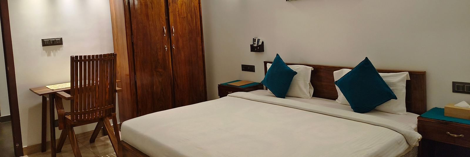 Wide view of a double bed with blue pillows, a chair and a cupboard in 102 room at Ananda Holiday Home, Wayanad 9