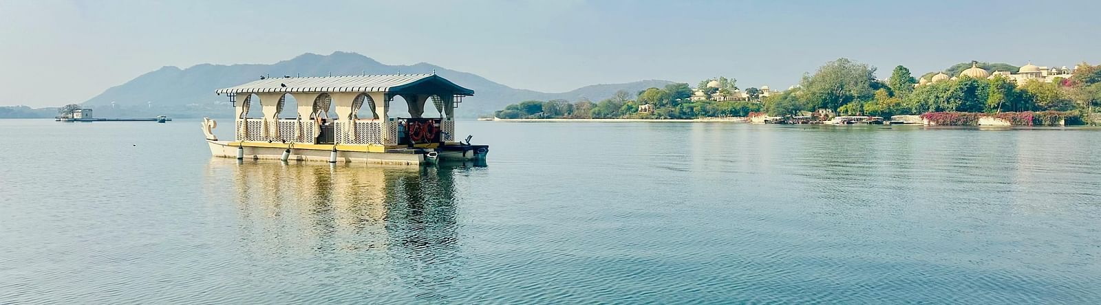 A traditional houseboat floats peacefully on the still, blue waters of a lake, with distant green hills under a clear sky.