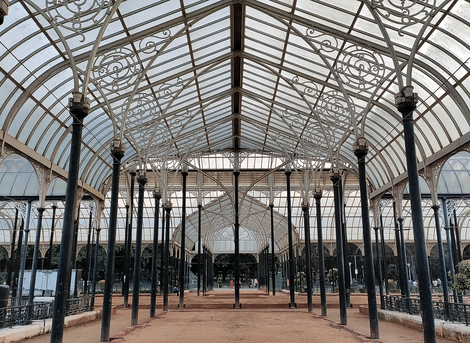 A spacious glasshouse with an ornate metal framework and tall, dark pillars over a sandy ground.