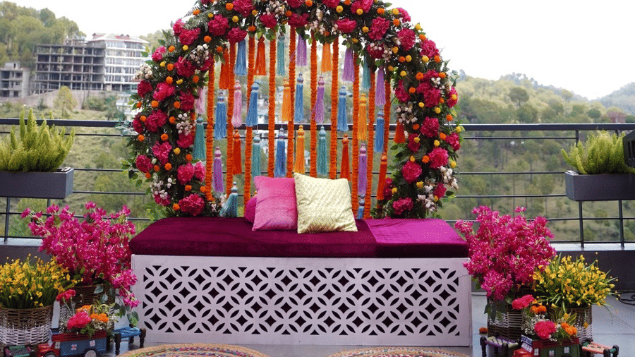 Colourful floral seating backdrop with marigold hangings, cushions, and decorative rugs set on an outdoor terrace | Ambrosia Sarovar Portico, Haridwar