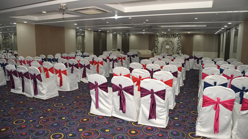 A spacious conference or wedding hall at Ambar Sarovar Portico, Gandhidham is arranged with multiple rows of chairs decorated with ribbons.