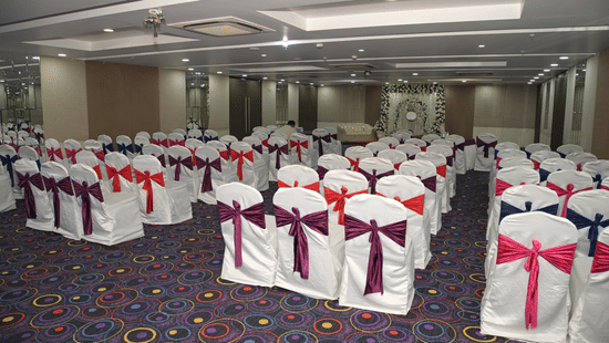 A spacious conference or wedding hall at Ambar Sarovar Portico, Gandhidham is arranged with multiple rows of chairs decorated with ribbons.