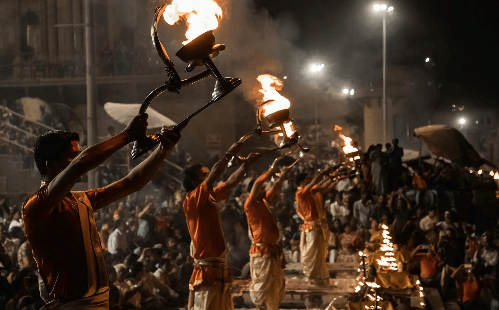 A side-angle image captures the scene of people performing Aarti at the bank of the Ganga River, highlighting the reverence and devotion as they hold lamps and sing prayers, with the shimmering water and evening lights creating a serene backdrop.