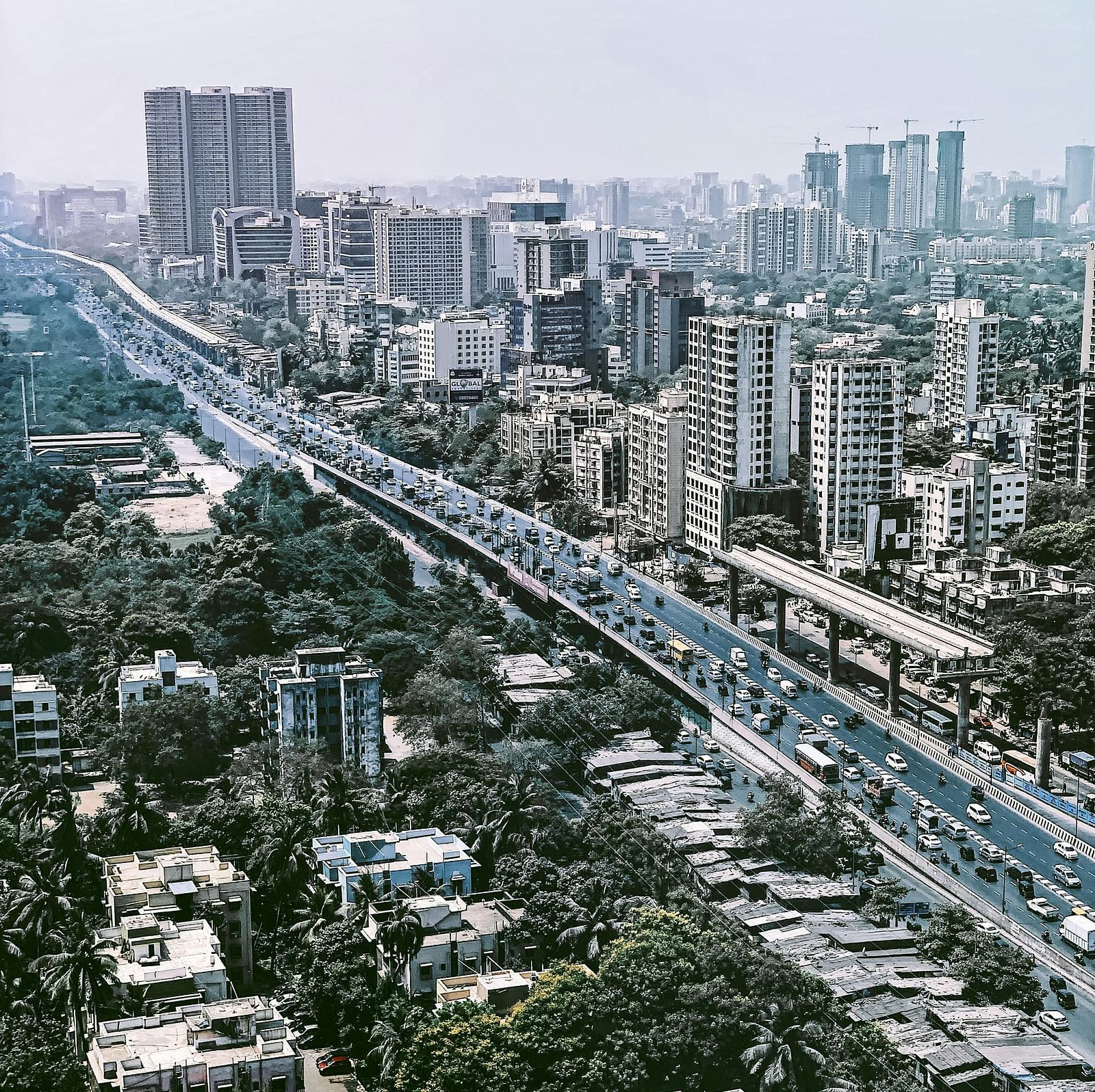 An aerial view of the city, with tall buildings on one side and buildings surrounded by trees on the other, separated by a road in between.