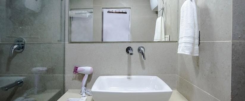 This image shows a modern hotel bathroom with a sleek, white rectangular sink on a beige countertop, a mounted mirror, and neatly arranged towels and toiletries | DLS La Serene Manali