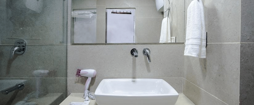 This image shows a modern hotel bathroom with a sleek, white rectangular sink on a beige counter top, a mounted mirror, and neatly arranged towels and toiletries | DLS La Serene Manali