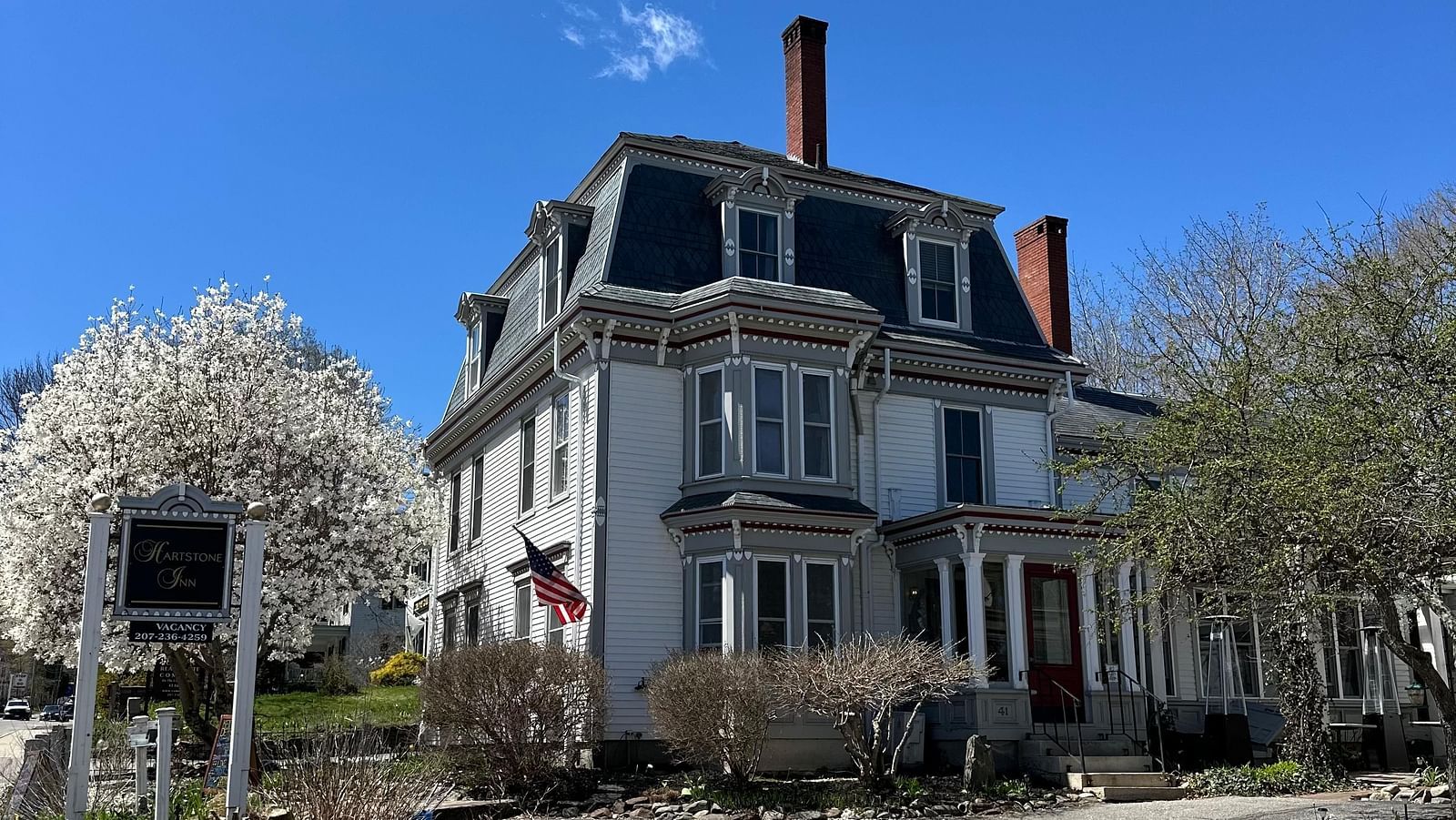Hartstone Inn on a sunny day, showcasing its grand Victorian architecture, American flag on top, and blooming trees lining the driveway.