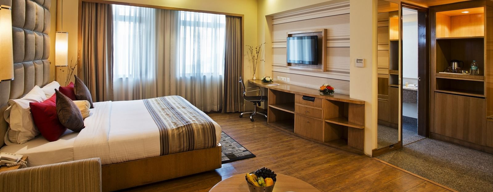 A sideview of a bed in a room with TV and a huge glass window | The Manor Bareilly