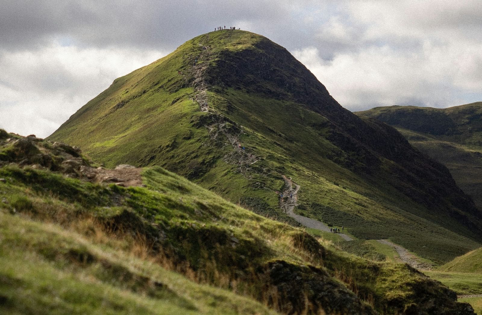 A steep, lush green mountain with a distinctive ridge and a rocky peak, set against a dramatic, cloudy sky.