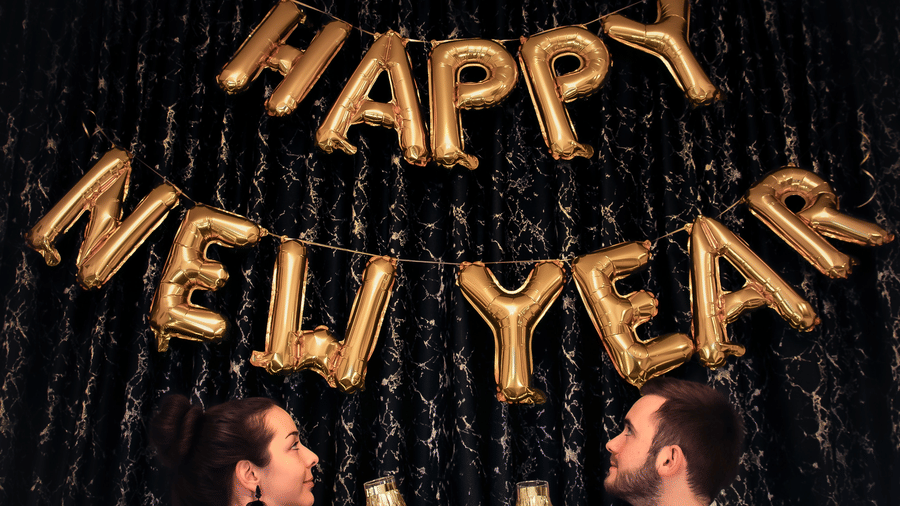 A couple holding drinks with happy new year made out of ballons on the wall