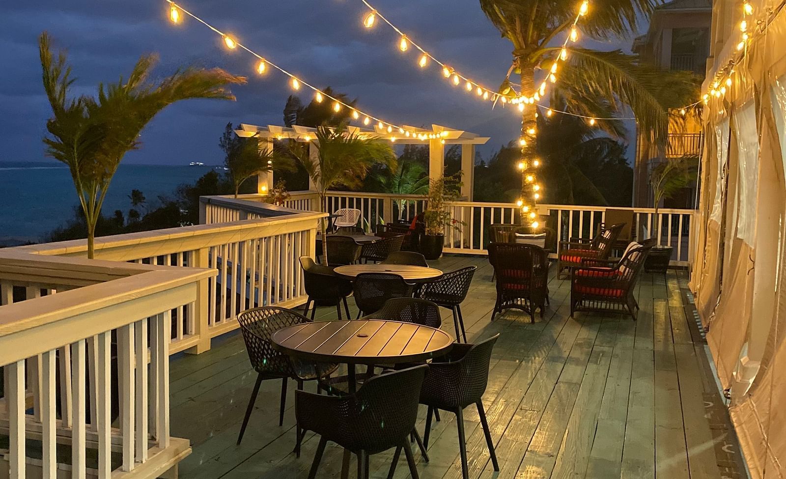 Outdoor dining area with string lights at dusk, white railing and palm trees in view | A Stone's Throw Away
