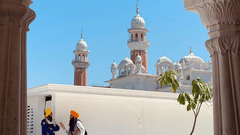 An arched doorway framing a view of a white temple with domes and minarets, with two people walking inside the temple complex, set against a bright blue sky.
