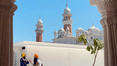 An arched doorway framing a view of a white temple with domes and minarets, with two people walking inside the temple complex, set against a bright blue sky.