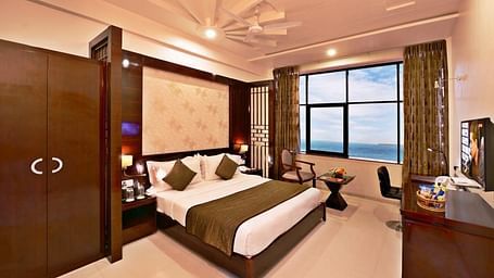 A cosy hotel room includes a king size bed with patterned bedding, a seating area near a window with a view, and warm lighting.