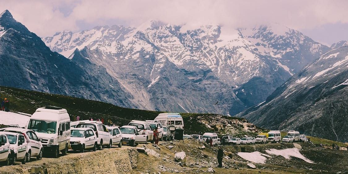Snow-dusted mountain pass with traffic and parked vehicles, set against a dramatic backdrop of rugged Himalayan.