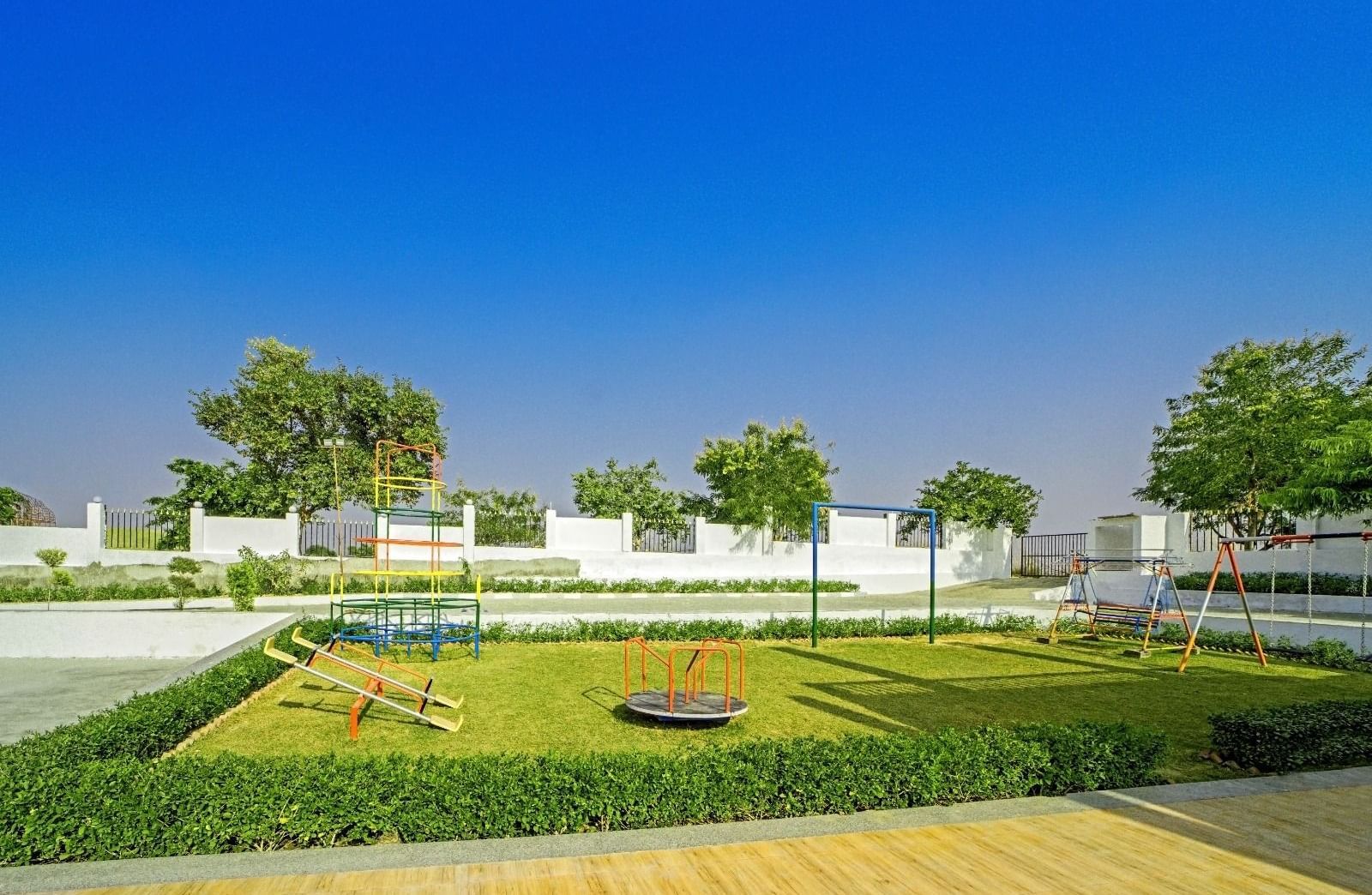 Children’s playground at Grassfield Riveria Resort, Jaipur, featuring swings, a roundabout, and lush greenery under a clear blue sky.
