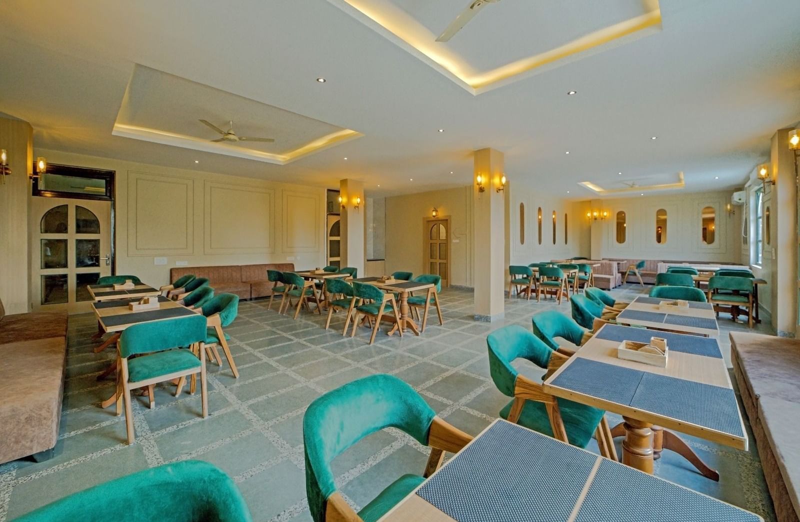 Elegant dining area at Grassfield Riveria Resort, Jaipur, with teal upholstered chairs, wooden tables, and warm lighting.