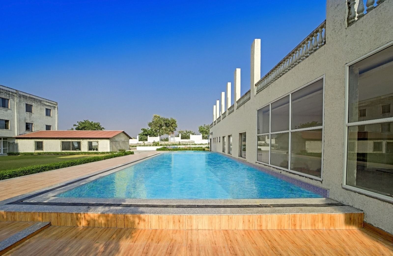 Outdoor swimming pool at Grassfield Riveria Resort, Jaipur, surrounded by modern buildings and a wooden deck area.