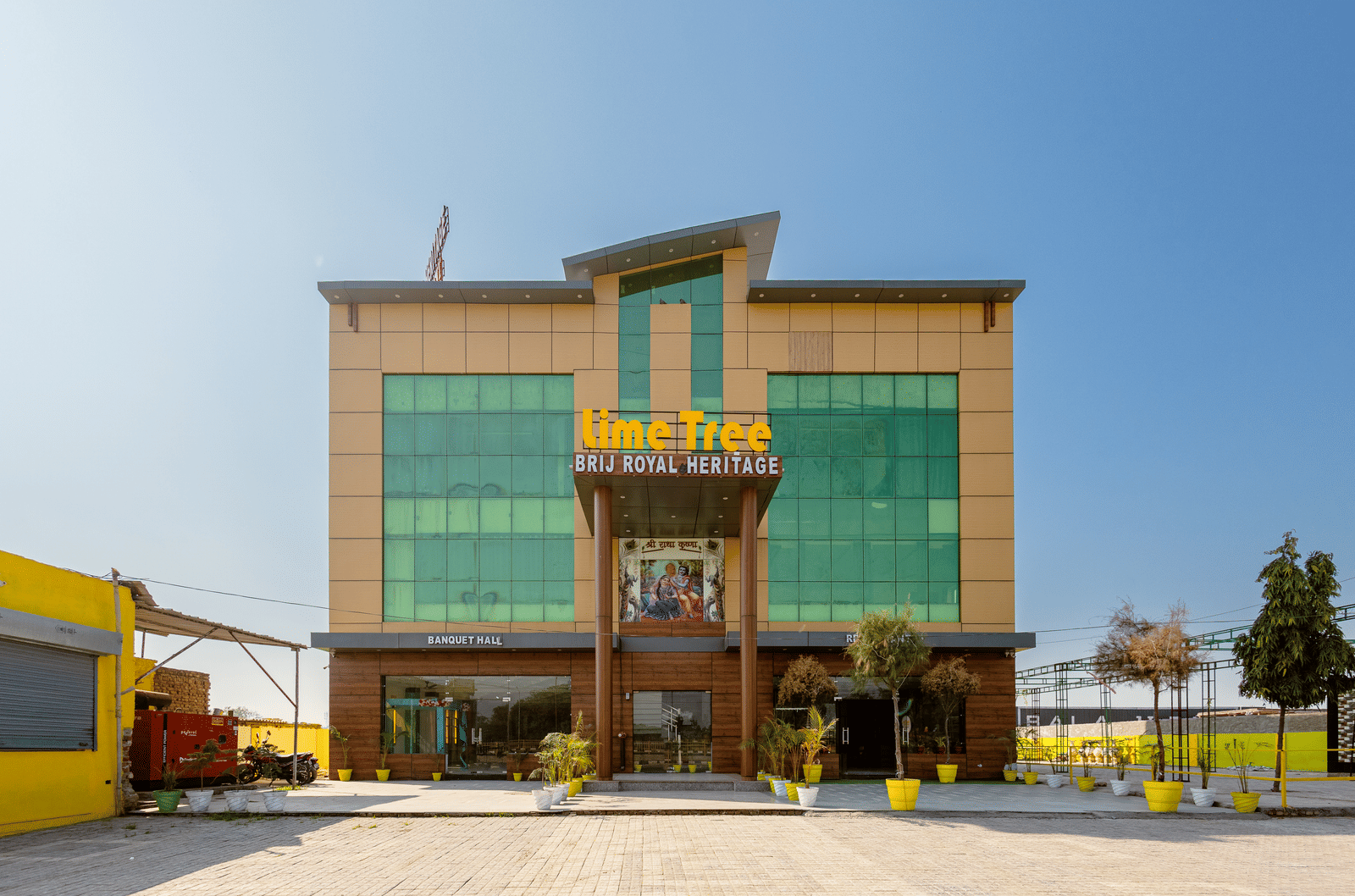 A daytime view of Lime Tree Hotels & Banquet, Vrindavan building with glass panels, yellow accents, and an open parking area in front.