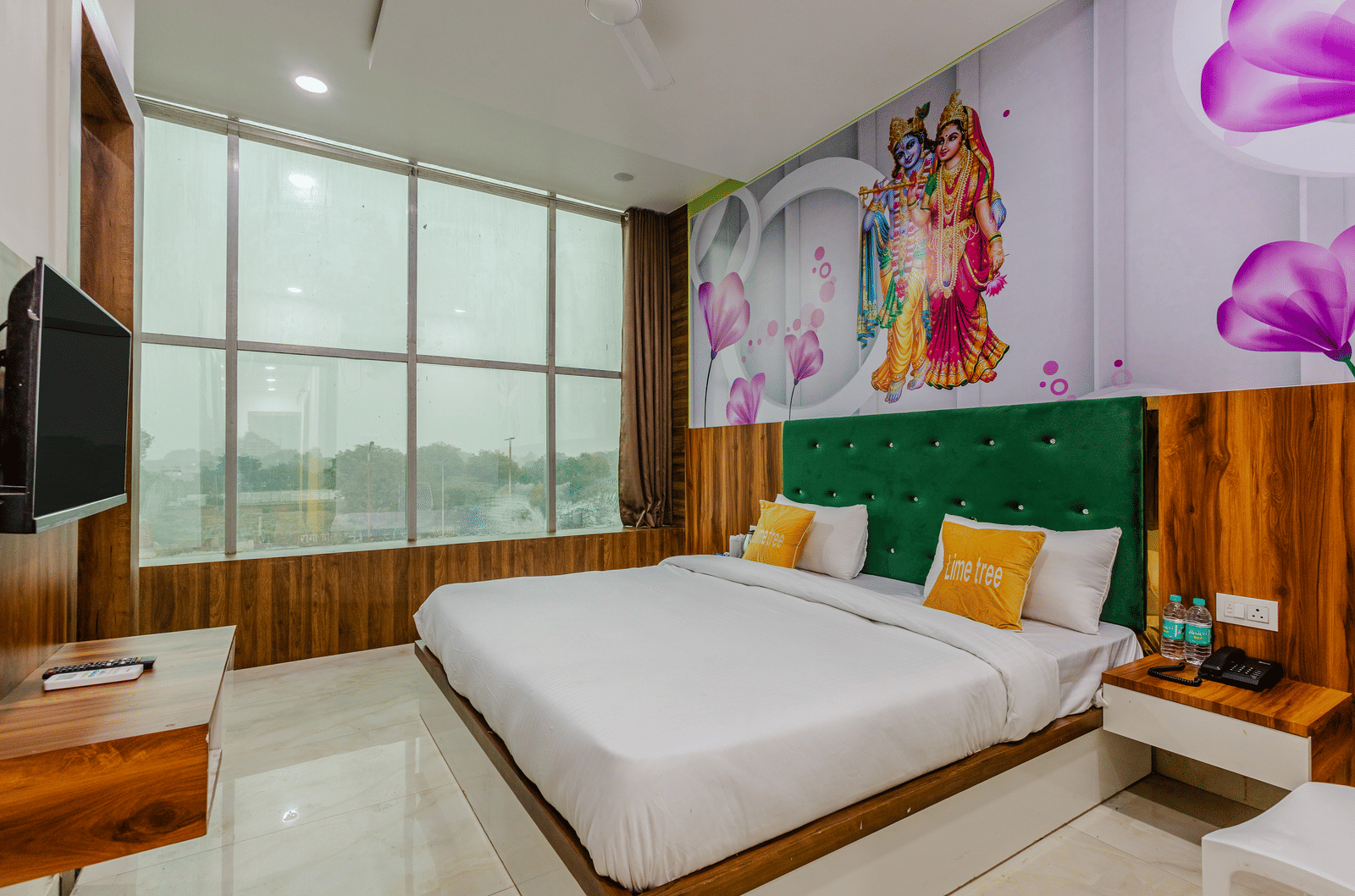 A bedroom with a green headboard, artwork above the bed, large windows, and a wall-mounted television - Lime Tree Hotels & Banquet, Vrindavan