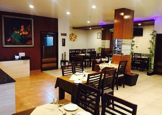 An elegantly set dining area with wooden chairs and tables, under ambient lighting and a painting on the wall - TGI Apple Inn, Jaipur 