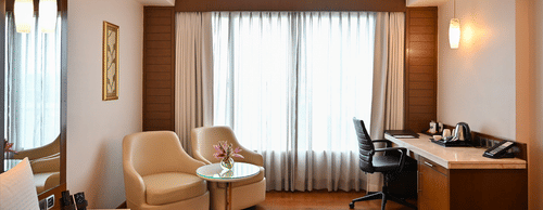 A spacious hotel suite featuring a seating area, desk, and large windows, offering a comfortable stay.