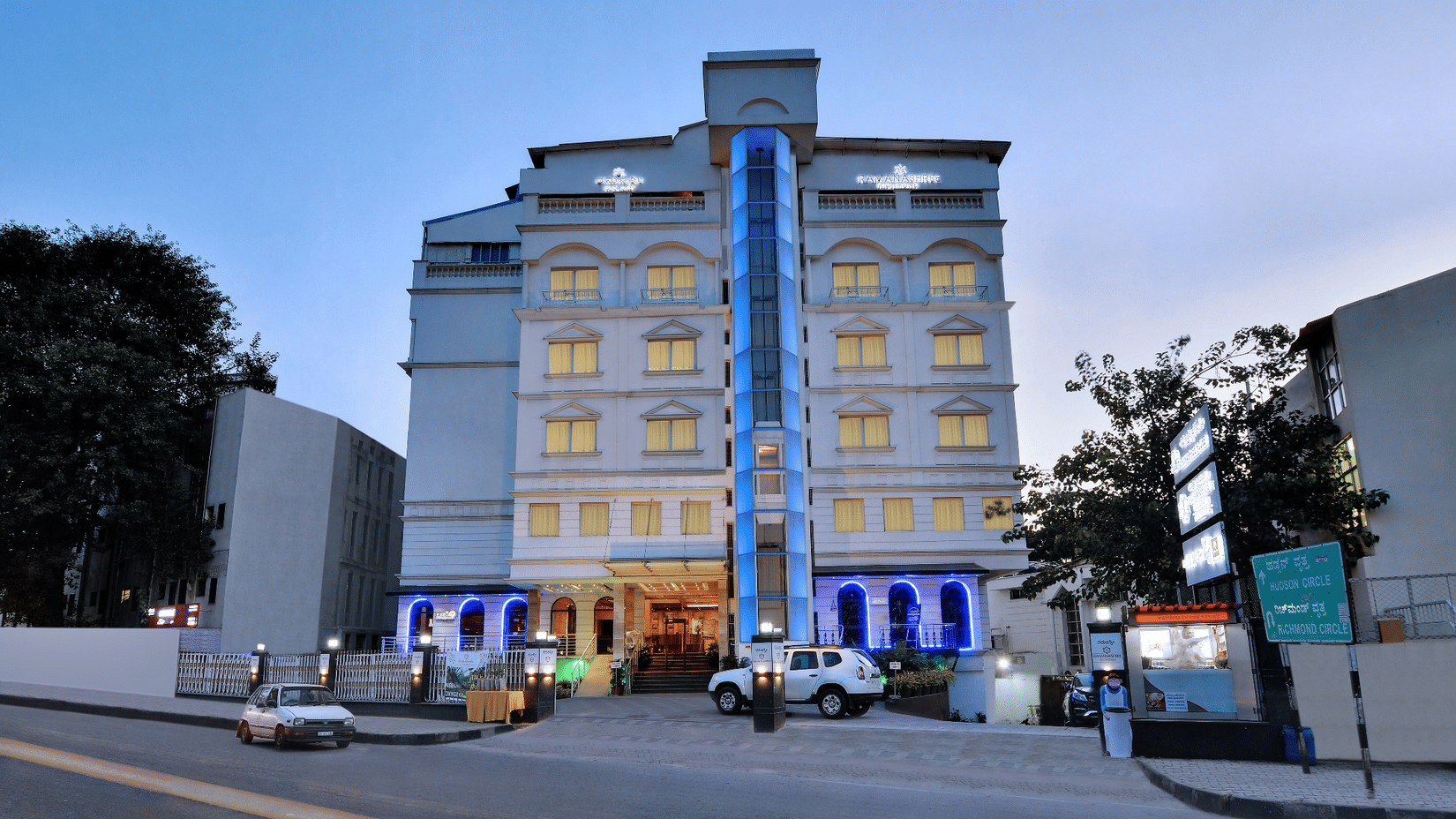 Hotel building exterior with entrance, parked cars, and lights in the evening.