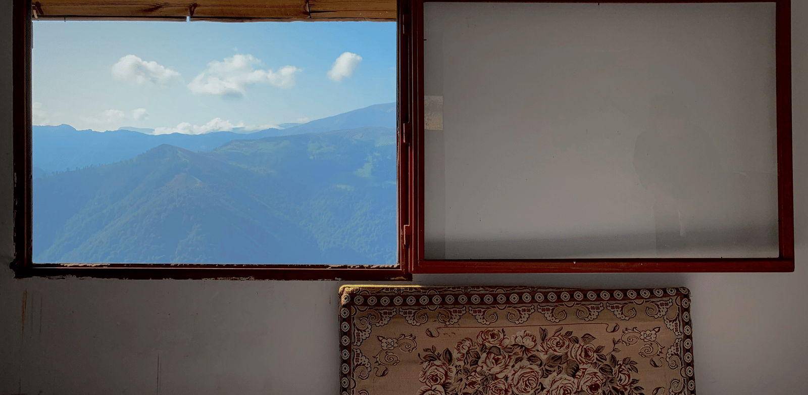 An interior wall with a view of green mountains and blue sky through an open window pane, next to a blank window and a rug below.