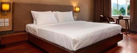 A double bedroom with sockets near the bed, two lamps on either sides of the bed, and a seating area near the glass window at The Hamuse Hotel, Kodaikanal