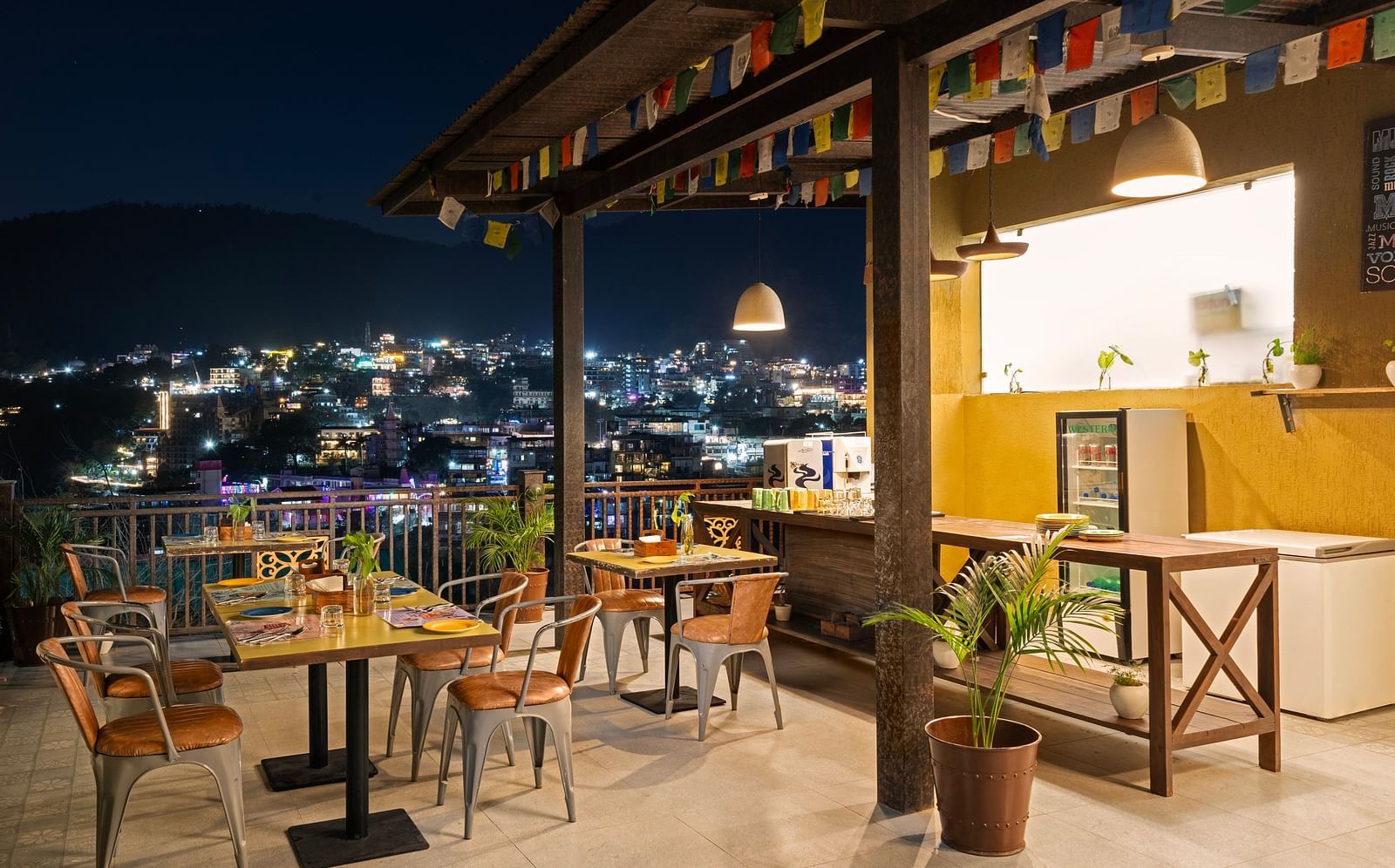 Panoramic view of city skyline from rooftop restaurant, with tables, chairs, and city lights at night at Bedzzz, Rishikesh 