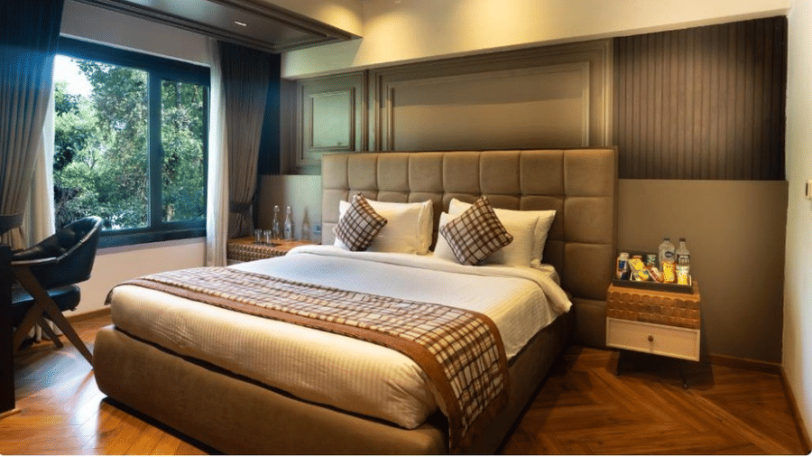 A bedroom featuring a large bed with a padded headboard, wooden ceiling beams, and a window overlooking green trees | Lawrence Solitaire Resort, Nainital