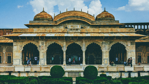 Facade of an ancient building with with intricate architectural design featuring lush garden and tourists, under a blue sky.