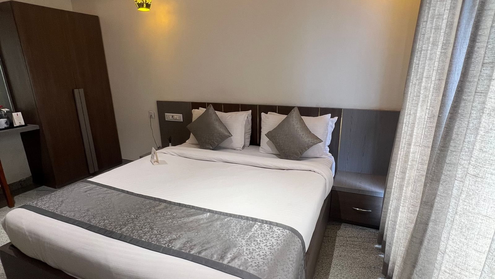 Corner view of Daksh Comfort room at Daksh The Valley Resort, Saputara, featuring a cosy room with a plush king size bed, a wardrobe, warm overhead lighting, and long closed curtains on one side of the room.